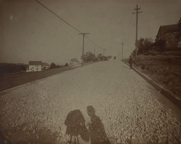 Perspective view of an unidentified street paved with gravel, with the photographer's silhouette. There is a house on a hill on the right, and more houses on the lower part of the hill on the left. A man is standing near the curb on the right halfway up the hill, and at the top of the hill in the distance is a man driving what may be grading equipment for road building. An inscription on the back of the photograph notes that Joseph Schubert may be the photographer. Joseph Schubert was a cabinet maker turned photographer in the mid-1800s. Joseph C. Schubert took over his father's photography business and in 1906 was elected mayor of Madison.