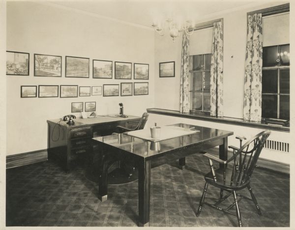 Interior view of Stanley Hanks' real estate office at 311 State Street. There is a chair in front of a desk with a calendar and two telephones. Above the desk on the wall are many framed photographs of Madison scenes. In the foreground is a work table and chair. The floor is carpeted and the two windows are framed by curtains.