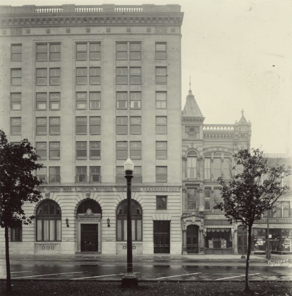 View across street towards the First National Bank and Central Wisconsin Trust Company on the corner of East Washington Avenue and S. Pinckney Street. The cornerstone for the First Central Building was laid in 1921. It housed these two businesses, plus others on multiple floors. This building later became the 'glass bank,' the First National Bank on S. Pinckney. On the far right is the Tittle Meat Market.