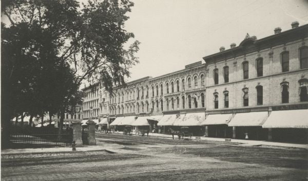 View across street towards the buildings on South Pinckney Street. The fence surrounding Capitol Park is on the left. The businesses have signs in their windows for boots and shoes; hardware, including paints, oils and glass; pianos and organs; drugs and medicines; and clothing.