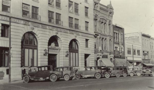 View from street of the First National Bank and Central Wisconsin Trust Company in the 100 block of South Pinckney Street. Other businesses include the Chop Suey Restaurant and hardware store on the right towards the end of the block.