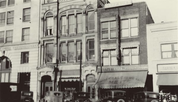 View from across street of the 100 block of South Pinckney Street between 1925 and 1929. The State Bank, Lucien S. Banks, President, is in the center. To the left is a partial view of the First National Bank and Central Wisconsin Trust Company. On the right of the State Bank is a Wholesale and Retail Meats store, the Chop Suey Restaurant, and the Kresge Company store.