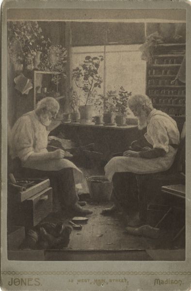 Carte-de-visite with a photograph of a painting by James R. Stuart. It depicts two white-haired, bearded shoemakers, with Langford on the left and his partner on the right. They are sitting on workbenches in front of a window with numerous flower pots. Each man is wearing an apron, and white shirts with rolled up sleeves. They are looking down at their work. There are boots and tools scattered on the floor in the foreground. On the right wall, next to the window, are shelves full of footwear. Stanley Hanks has annotated the verso of the photograph: "Given me by H.E. Shaw. The shop was at 17 N. Pinckney." In a separate hand is the notation: "For W. DeLamaytn." The date 10/25/40 refers to the date Stanley Hanks acquired the photograph.