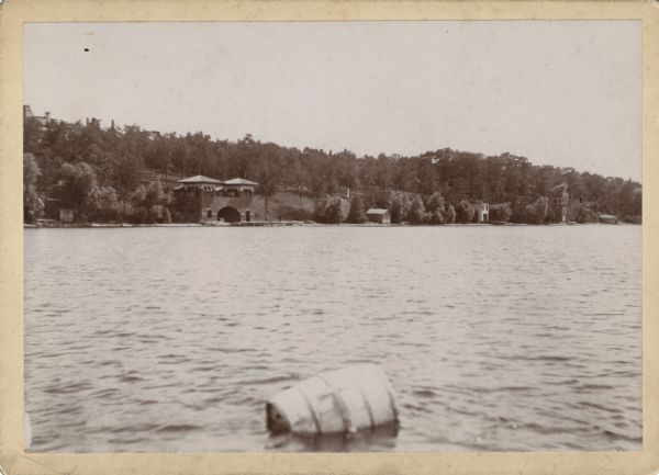View from lake towards the Frank Lloyd Wright Boathouse, also known as the City Boat House. The boathouse was at the foot of North Carroll Street on Lake Mendota. It was an early design by Frank Lloyd Wright. There is a barrel floating in the lake in the foreground. 
