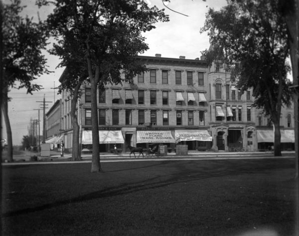 View across Capital lawn towards the corner building at 100 S. Pinckney, on the Brown or Bruen Block. The stores, from the left, are Sullivan & Heim, Blasius Pianos (the awning says Groves Piano Ware Rooms, J.W. Groves, Mgr.) and an awning that reads: "Dry Goods Clearance Sale of Shirt Waists." To the right of the clearance sale is the State Bank. There is a woman walking past the Sullivan and Heim building; a horse and buggy is moving down the street, piano packing cases are in front of the piano store, and several bicycles are parked in front of the State Bank.