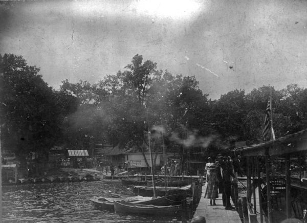 View from pier of groups of people walking along a Lake Monona dock and on the walkway to the shore. On the right is an excursion boat with a United States flag. Sailboats and rowboats are along tethered on the left side of the dock. On the shore is a building with a sign advertising an "Excursion to the Dells." Behind the fence a canvas awning provides entrance to the excursion area.