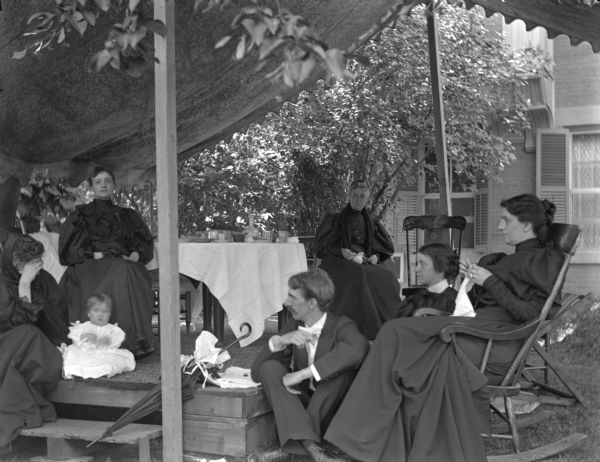 An informal group is seated under an awning in the garden of the Fairchild house at 302 Monona Avenue (later renamed Martin Luther King Boulevard). Pictured are Mrs. Fairchild, Mary Fairchild Morris, Sallie Fairchild Bacon, Lucien M. Hanks and others. This garden party photograph is one of a series. The table in the background is set with dishes, napkins, cups and glasses.<p>The house was constructed in 1850 at 302 South Wisconsin Avenue (renamed Monona Avenue in 1877) near Lake Monona, by Jarius Fairchild  and was later substantially altered with additions. After the Civil War, the house became the home of Fairchild's son, Lucius, Civil War hero, Wisconsin governor (1866-72) and foreign diplomat. During his six years as governor, the Fairchild House was the state's executive residence, as no such property was then owned by the state. The residence was therefore a focus of Madison's political and social life during the last half of the 19th century.</p>
