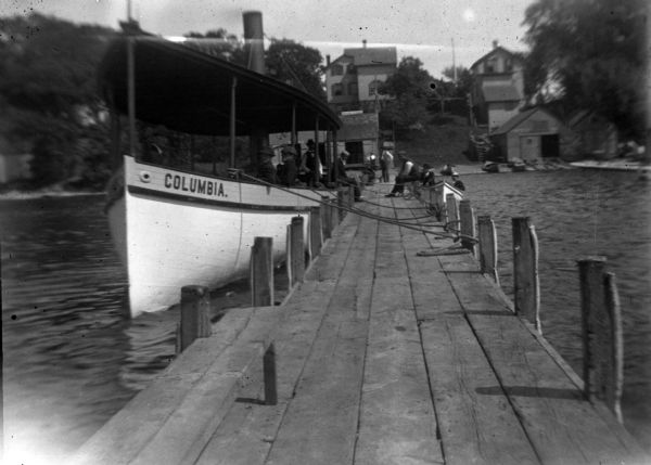 View down pier towards the "Columbia" steamboat at the boat landing. The shoreline is the the background. There are three or four people on the steamboat and another three or four hanging out on the dock. Two men are in a dinghy on the right.

Charles Bernard built the "Columbia" in 1893, the second of three steamers that his family ran as two hour excursion boats. Bernard's Boathouse was located at 622 East Gorham Street on Lake Mendota.