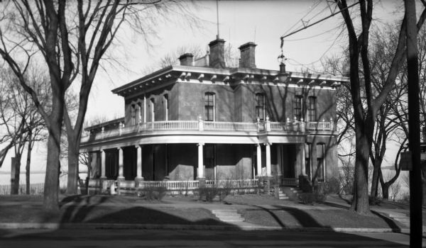 View across street towards the house at 130 East Gilman Street, which was built between 1854-1856. Lake Mendota is in the background. The stone building features a wrap-around porch and a balcony. 

Soon after Governor Jeremiah Rusk bought the home, he sold it to the State of Wisconsin. From about 1885 until 1950 this house served as home to 18 Wisconsin governors.