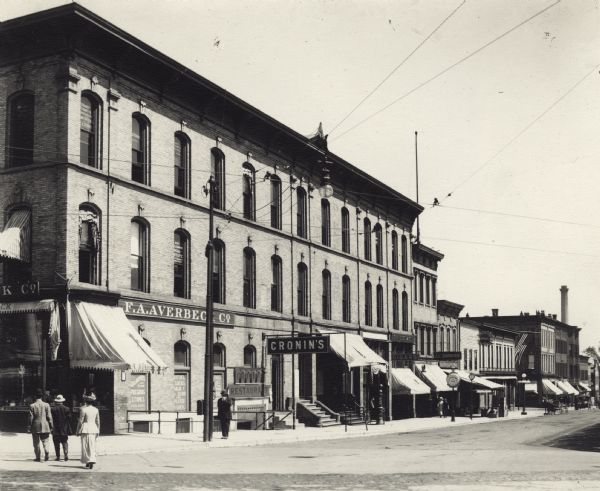 View across street towards the F.A. Averbeck Company, a jewelry and clock shop, and Cronin's Restaurant, on the corner of Main and South Pinckney Streets. Pedestrians are walking on the street and on the sidewalks. On the far right are horse-drawn carriages and automobiles. This corner later became the site of the Tenney Building.
