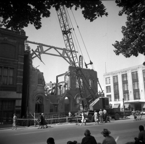 View across Mifflin Street, towards Madison City Hall as it is being demolished. On the street women are walking around the construction to get to the sidewalk. Men in work clothes are standing in the street near a crane. In the foreground men are sitting and watching the demolition.