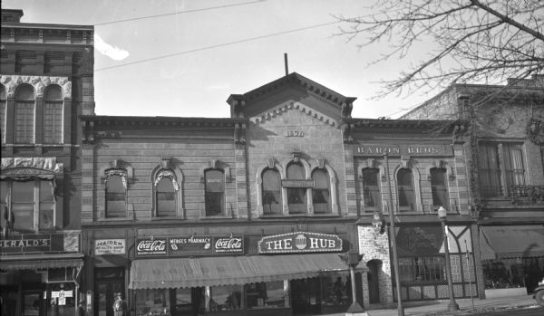 View from across street of the facade of the Kronke building on West Mifflin Street. From the left, businesses are the Maiden Beauty Shop, Menges Pharmacy, The Hub, the Heidelberg Hofbrau, and Baron Bros. Inc. There is also a dentist's office on the second floor. The Kronke building was built in 1871.