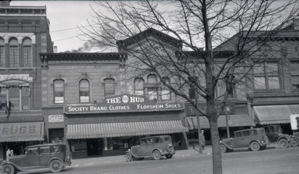 View across street towards The Hub, a men's clothing store. Above the striped awning is a sign reading: "Society Brand Clothes, Florsheim Shoes." Directly to the left is the door to the Maiden Beauty Shop, and the Menges Pharmacy. Above the pharmacy is Buckmaster Jeweler. On the second floor, next to Maiden Beauty Shop is R.W. Niederer, Dentist. On the right side of The Hub is a partially obscured shop that may say "Universal." There are automobiles angle parked on the 100 block of W. Mifflin Street.