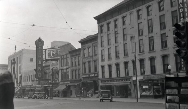 View across street towards storefronts on the first block of East Mifflin Street. In the center is Conklin and Sons, an ice and heating fuel provider in Madison. To the left is the Strand Theater, which is showing Charles Farrell's "Heartbreak." On the right is Woldenberg's, a women's clothing shop, and Walgreen Drugs. There is a newsstand in the right foreground below a traffic light.