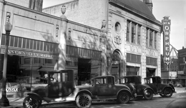 View across street towards 116-118 N. Fairchild Street towards a row of 1920s automobiles angle parked outside Esser's City Market. To the right of the market, across State Street, is the Orpheum Theatre.
