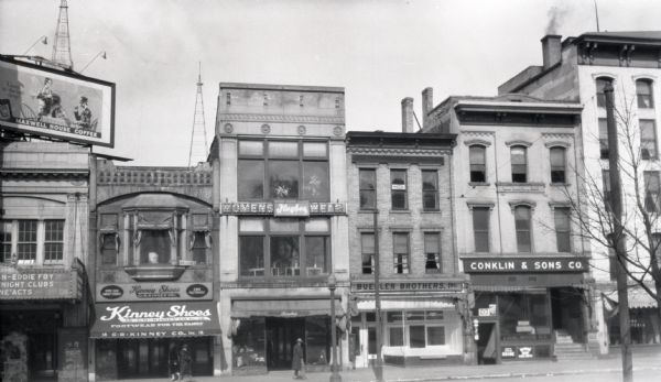 View from across street towards the first block of East Mifflin Street. The businesses are, from left to right: The Strand Theater, Kinney Shoes, Hughes Womens Wear, Buehler Brothers, and Conklin and Sons Co. On the far left on top of the Strand is a billboard for Maxwell House coffee.