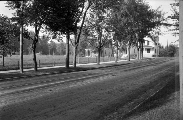 View of Baldwin Street in the mid 1920s from Johnson Street, which is unpaved, and has what appear to be streetcar tracks. There is a line of trees on Johnson Street partially obscuring three houses in a row on Baldwin Street. There is a low fence on the far side of the sidewalk surrounding an empty field.