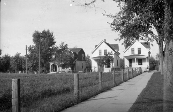 Looking east down sidewalk running along East Johnson street towards three houses standing in a row on North Baldwin Street in the mid 1920s. On the right is a line of trees along the terrace, and on the left is a fence surrounding a field. On the sidewalk at the corner is a young child sitting on a wagon.  The street has been widened and the only house still standing is at 305 North Baldwin Street.