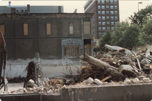View from Wisconsin Avenue towards the razing of Manchester's Department Store at 14 E. Mifflin Street. The foundation is exposed. A man wearing a hardhat is standing on the right. The brick wall on the building next door has a newly revealed sign for "Collyer's Drugs / Soda."