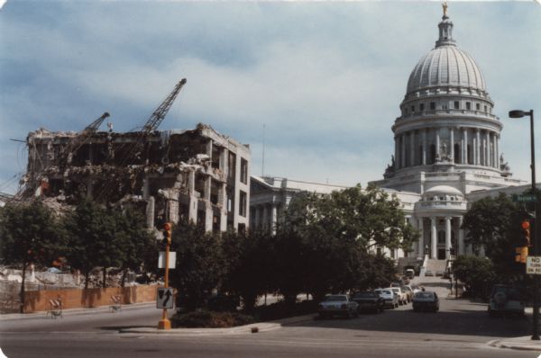 View up Wisconsin Avenue at West Dayton Street towards the razing of Manchester's Department Store. There are construction workers working near two cranes set up on the lot. The Wisconsin State Capitol is in the background.