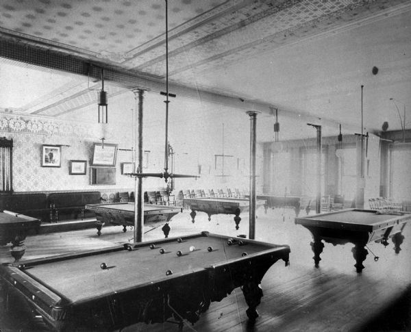 Six billiard tables in a large room with high ceilings at the Park Hotel on Carroll Street. Benches and chairs line the back wall, and large windows are along the wall on the right.