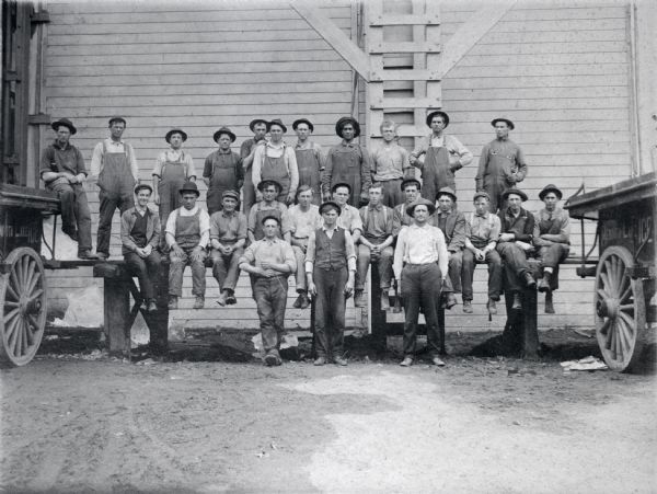 Outdoor group portrait of Conklin Ice House employees posing on a loading dock in front of the ice house on Lake Mendota. Wagons are parked on the left and the right.