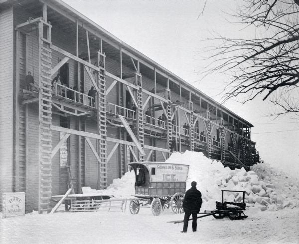 Conklin Ice House on Lake Mendota. A man is standing in the snow-covered yard in the foreground near a sleigh, and a carriage with a sign painted on the side reading: "Conklin & Sons Mendota Lake Ice." Behind the carriage is a large pile of ice and snow. Men are working on the second floor gallery near wooden ladders and pulleys. On the far left is a box with painted with the words "Howe U.S. Standard," probably a scale.