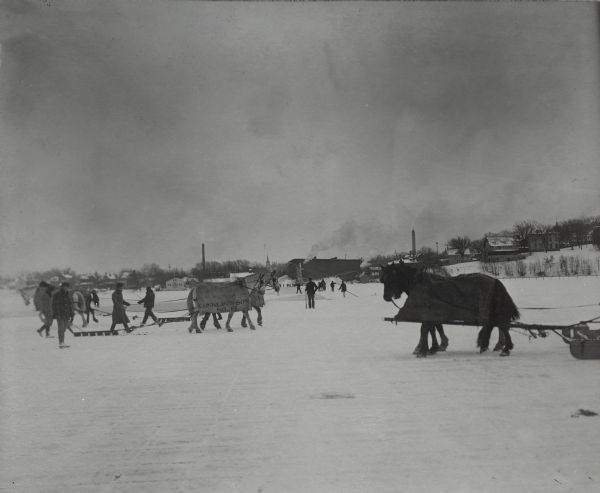 View across ice towards a group of men, employees of the Conklin Ice House, which is in the distance, guiding horses pulling ice cutters. The horses are wearing blankets. In the foreground, grid lines are visible in the ice where the ice cutters have passed. The blanket on the horse in the center reads: "Conklin and Sons N. 15." In the background are a number of men carrying long poles. In the far background on the right is a water tower.