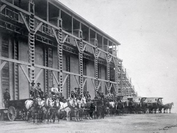 Outdoor group portrait of men posing on horse-drawn wagons parked in front of the Conklin Ice House. In the background on the right is Lake Mendota.