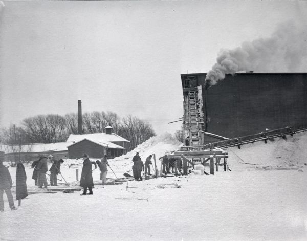 View across ice on Lake Mendota towards men using long poles to maneuver ice up to the conveyor belt to the Conklin Ice House.