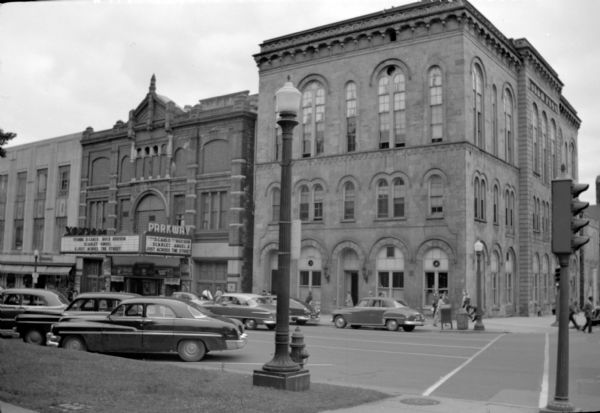 Exterior view from sidewalk of City Hall, located at the intersection of West Mifflin Street and Wisconsin Avenue. The Parkway Theatre, featuring the films "Scarlet Angel" and "Just Across the Street" is next door on the left. Automobiles are parked at an angle along the curb.