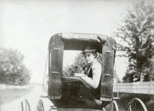 An unidentified woman posing while sitting and looking through the open back of a carriage on a road.