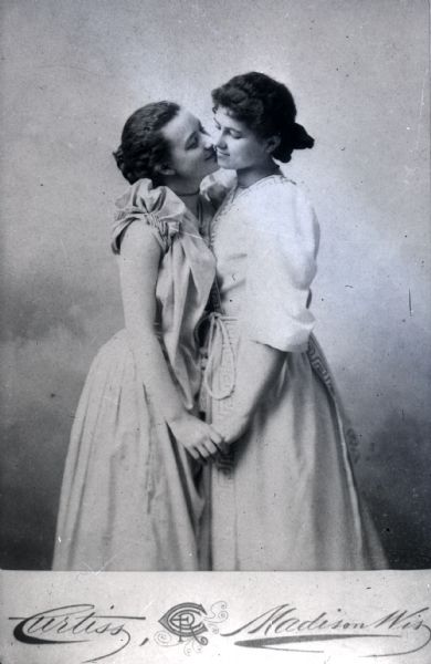 Three-quarter length cartes-de-visite portrait of two women posing for a portrait in front of a painted backdrop. They are facing each other and holding hands.