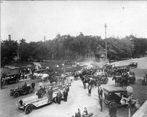 Elevated view of motorized fire trucks, horse-drawn fire trucks, automobiles and water wagons gathered on East Washington Avenue. All the vehicles are accompanied by firemen, town officials and townspeople. Houses are along the corner of the side street in the background. There is an empty lot on the left, and a brick building behind it with a sign that reads, in part: "Garage, — Day and Night."