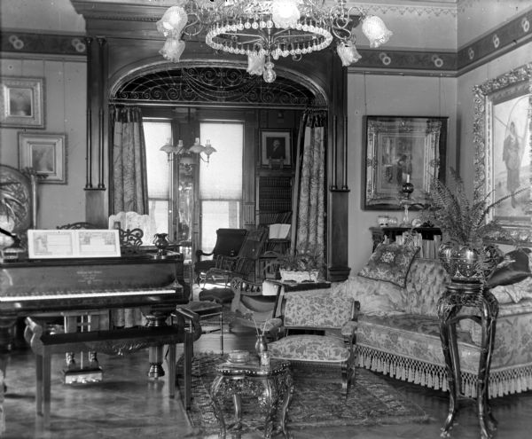 An interior view of a sumptuous Victorian parlor, with a Steinway piano, chandelier and ferns at the William Vilas house, 12 E. Gilman Street. A curtained archway leads to another room with windows on the far wall.