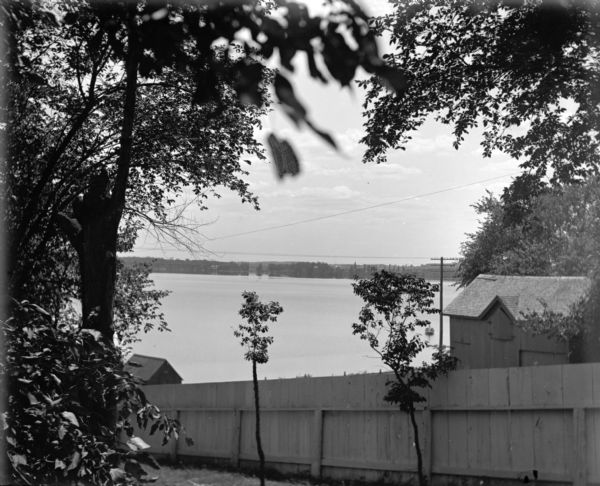 View down hill towards a board fence and two outbuildings on the shore of Lake Monona, taken from the back yard of the Lucius Fairchild home at 302 Monona Avenue (later renamed Martin Luther King Boulevard). The far shoreline is in the distance.