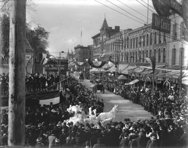 Elevated view of a white horse and a white float rounding the corner in a parade going down South Pinckney Street at Main Street. Other carriages and floats are proceeding down the street into the distance. A large crowd is watching from the sidewalks. There is a band playing on a stage on the corner of the Capitol Square Park on the left.
