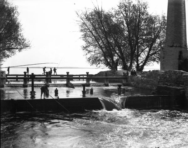 View across the Yahara River towards a tall smokestack on the opposite shoreline, with Lake Mendota in the background. Remnants of the foundation and supports of what had been Farwell's Madison Mills are seen in and along the river. Men and children are fishing and playing in the river all along the dam. In the background is the far shoreline of Lake Mendota.