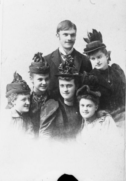 Group portrait of six young people. The five women are wearing hats, and are posed in front of young man wearing a suit.