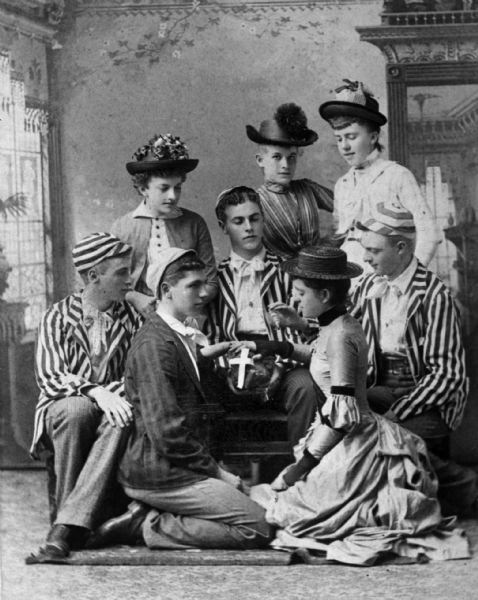 Group portrait of four men and four women posing while sitting and standing in front of a painted backdrop. The men are wearing striped jackets, and two of the men are wearing striped caps. The women are wearing summer hats and dresses. One of the men and one of the women are kneeling and facing each other in front of the man sitting in the center, who is holding an object in his hands covered with a white cross.