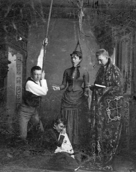 Studio portrait in front of a painted backdrop of two men and two women in a dramatic pose. The woman in the center has her head in a noose and is sticking out her tongue as she clenches her fists. A man on the left is pulling on the rope as if to hang the woman. On the right side, another man is reading from a book and wearing a heavy carpet as a robe. A woman is kneeling on the floor wiping her eyes with a cloth.