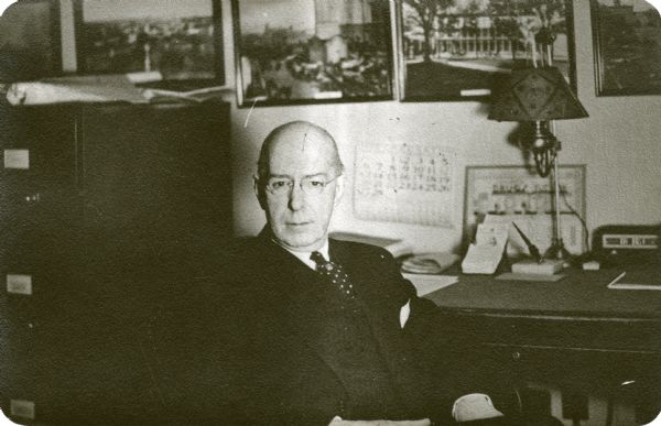Stanley Hanks gazing at the camera while sitting in his office at 24 West Mifflin Street. There are framed photographs on the wall behind him of businesses and residences in Madison. On his desk is a pen, a lamp and a radio.