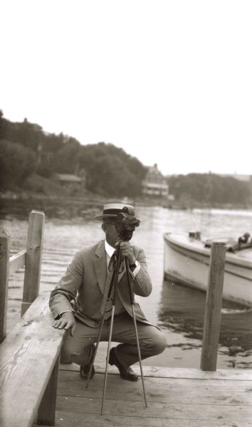 A photographer is crouching on a wooden pier behind a small camera on a tripod to film C.B. (Chandler Burnell) Chapman, a realtor and good friend of Stanley Hanks, as he swam across Lake Mendota to Picnic Point from the foot of North Carroll Street. The swim took one hour and forty minutes. There is a shoreline in the background.

He first made that swim in 1886 with Stanley Hanks, Henry T. Sheldon, and Captain Tom Isabell.
