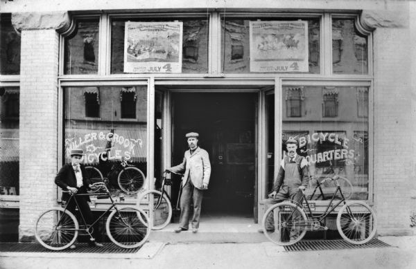 From left to right: Llewellyn Miller, George L. Crook and Ralph Miller all posing with bicycles and standing outside their bicycle shop at 121 King Street. The windows are reflecting the awnings and windows of the buildings across the street. There are large circus posters in the windows above the doorway of the bicycle shop.