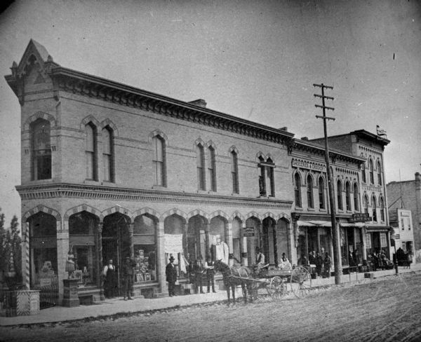 View across East Main Street towards a row of businesses, looking southeast. From the left, the corner building has a tobacco shop with a law office above, the Northwestern Telegraph Office, a shop for "Groceries and Crockery," and the Park Bakery. Beyond the Park Bakery is a storefront with a sign that reads: Furniture," and above is a large rocking chair attached the roof line. Groups of men are gathered on the sidewalk in front of the storefronts.