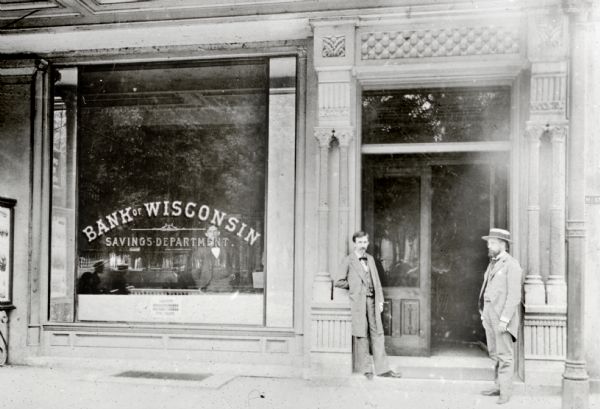 View from street towards two men posing at the entrance to the Bank of Wisconsin on Main Street. The window has Savings Department painted on it, and inside is a man standing wearing a suit with his hands in his pockets.