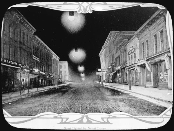 View down center of unpaved Main Street, lit by Nernst lamps. On the left side of the street is M.L. Nelson's office at 119. On the right side of the street is a cigar store, a trunk factory and a millinery, which is at 120 Main Street. 