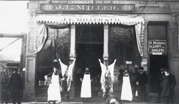 Outdoor group portrait of, left to right, William F. Fuller, James L. Miller, and Albert Zimmerman standing outside their shop at 24 W. Mifflin Street. In the center is a display of two gutted cows. To the right of the butcher store is the doorway to H. Nunns Pianos and Organs. There are several people standing on the street, including children.