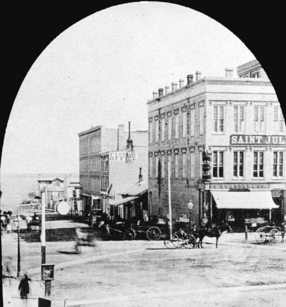 Elevated view of the corner of Pinckney and Main Streets, with men on horse-drawn wagons and pedestrians on the sidewalks. On the right corner is the S. Klauber and Company Dry Goods store, and above is a sign for "St. Julien Billiard Rooms." Further down Pinckney Street is the Patriot Office, and in the background down the hill is Lake Monona.