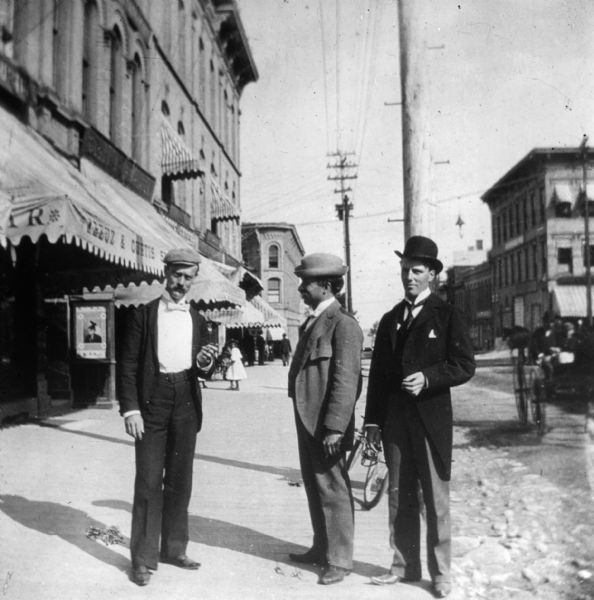 View of three men standing together on the board sidewalk outside Kreuz and Curtis Shoes, 21 S. Pinckney Street. The men are wearing suits, ties and hats, and all three men are smoking. Behind the man on the right is a bicycle is parked against a telegraph pole. There are people riding in a wagon on the unpaved street on the right.